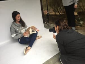 Behind the scenes shot of a pet photographer taking a photo of a woman with a small tan dog on her lap, sitting on a white backdrop set up at an animal shelter